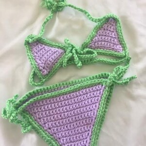Puede incluir: Un conjunto de bikini de ganchillo de dos piezas con copas triangulares de color lavanda y braguita a juego, ambos ribeteados con hilo verde lima brillante. La parte superior incluye lazos ajustables y la inferior tiene lazos laterales.