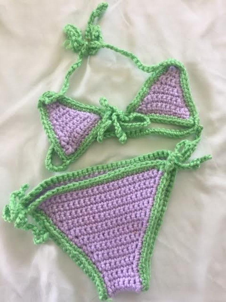 Puede incluir: Un conjunto de bikini de ganchillo de dos piezas con copas triangulares de color lavanda y braguita a juego, ambos ribeteados con hilo verde lima brillante. La parte superior incluye lazos ajustables y la inferior tiene lazos laterales.