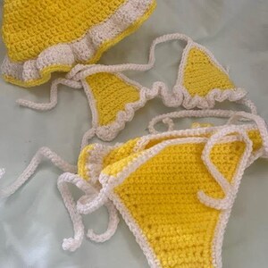 Puede incluir: Un conjunto de bikini de ganchillo amarillo brillante con un sombrero de pescador a juego. La parte superior e inferior del bikini presentan un borde de ganchillo blanco. El conjunto incluye largos lazos blancos para ajustar las piezas tejidas.
