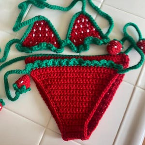 Puede incluir: Un conjunto de bikini de ganchillo de dos piezas con un patr&oacute;n de fresa roja, detalles de semillas blancas y ribetes verdes. La parte superior tiene copas triangulares con lazos verdes, y la parte inferior presenta una base roja con una cintura de volantes verde.