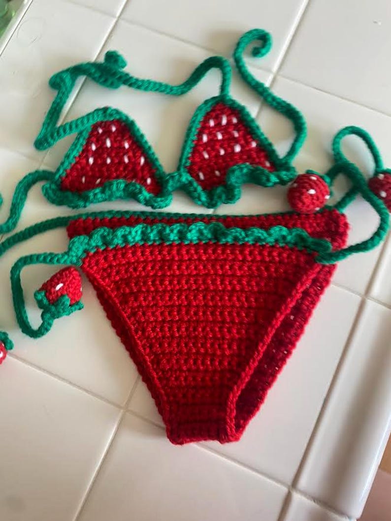 Puede incluir: Un conjunto de bikini de ganchillo de dos piezas con un patr&oacute;n de fresa roja, detalles de semillas blancas y ribetes verdes. La parte superior tiene copas triangulares con lazos verdes, y la parte inferior presenta una base roja con una cintura de volantes verde.