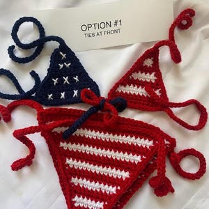 Puede incluir: Un conjunto de bikini de ganchillo que incluye un top triangular azul marino con estrellas blancas bordadas y un top triangular rojo con rayas horizontales blancas. La parte inferior es roja con rayas blancas y un lazo azul marino al frente. Texto: OPTION #1 TIES AT FRONT.