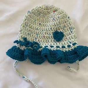 Puede incluir: Un sombrero de ganchillo hecho de hilo verde claro con motas multicolores, con un borde de volantes azul cerceta oscuro, una aplicaci&oacute;n de coraz&oacute;n de ganchillo azul cerceta, una banda tejida azul cerceta y dos lazos trenzados colgando.