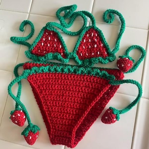 Puede incluir: Un conjunto de bikini de ganchillo en rojo y verde. La parte superior presenta dos copas triangulares con semillas bordadas en blanco y ribete festoneado verde. La parte inferior es roja con bordes festoneados verdes y dijes de fresa.