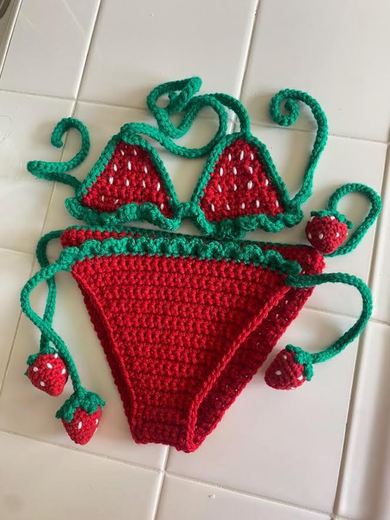 Puede incluir: Un conjunto de bikini de ganchillo en rojo y verde. La parte superior presenta dos copas triangulares con semillas bordadas en blanco y ribete festoneado verde. La parte inferior es roja con bordes festoneados verdes y dijes de fresa.