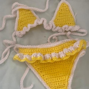 Puede incluir: Un conjunto de bikini de ganchillo de dos piezas con una base de hilo amarillo brillante y ribetes de color crema. La parte superior consta de dos copas triangulares con bordes con volantes y tiras largas. La braga es de corte cl&aacute;sico.
