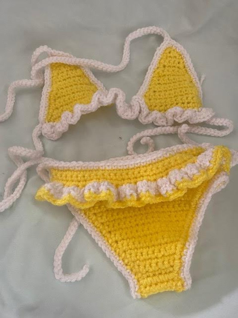 Puede incluir: Un conjunto de bikini de ganchillo de dos piezas con una base de hilo amarillo brillante y ribetes de color crema. La parte superior consta de dos copas triangulares con bordes con volantes y tiras largas. La braga es de corte cl&aacute;sico.