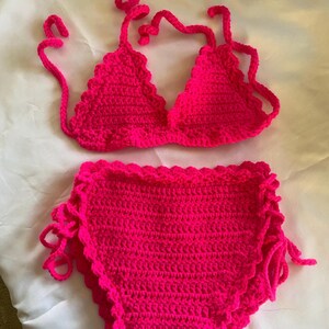 Puede incluir: Un conjunto de bikini de dos piezas tejido a ganchillo en color rosa intenso. La parte superior presenta copas triangulares con bordes festoneados y tiras finas para atar. La braga de talle alto incluye un borde festoneado y lazos laterales.