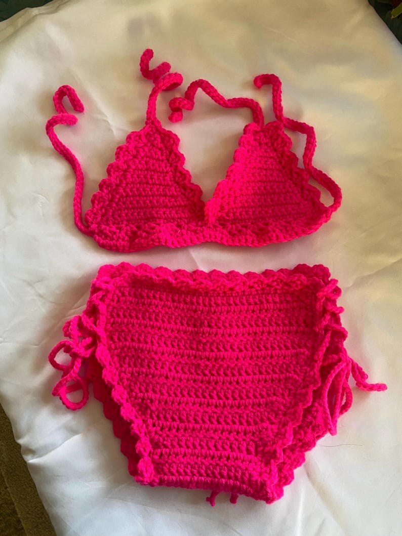 Puede incluir: Un conjunto de bikini de dos piezas tejido a ganchillo en color rosa intenso. La parte superior presenta copas triangulares con bordes festoneados y tiras finas para atar. La braga de talle alto incluye un borde festoneado y lazos laterales.