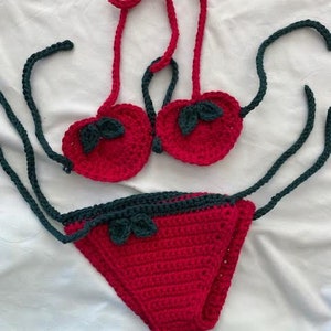 Puede incluir: Un conjunto de bikini de ganchillo de dos piezas en rojo brillante y verde oscuro. La parte superior presenta dos copas en forma de coraz&oacute;n con detalles de hojas verde oscuro y tiras rojas. La parte inferior es una pieza triangular roja.