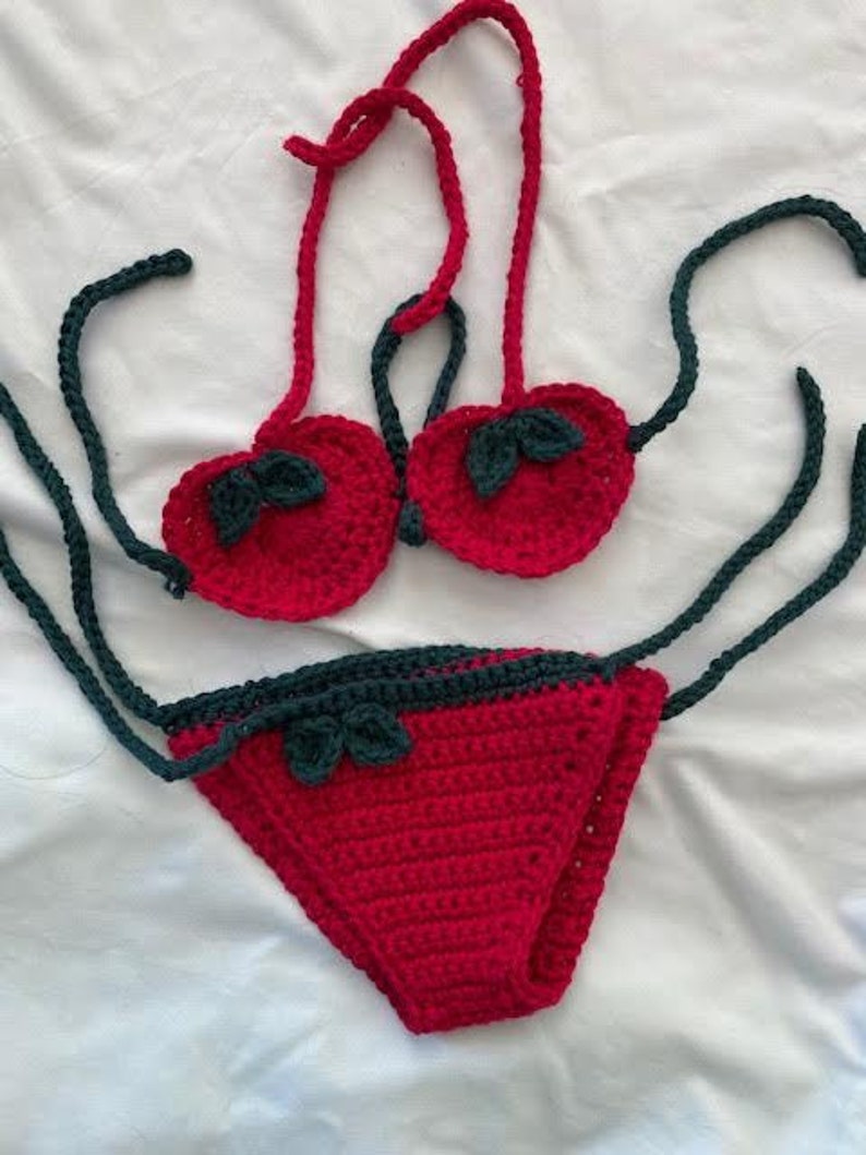 Puede incluir: Un conjunto de bikini de ganchillo de dos piezas en rojo brillante y verde oscuro. La parte superior presenta dos copas en forma de coraz&oacute;n con detalles de hojas verde oscuro y tiras rojas. La parte inferior es una pieza triangular roja.