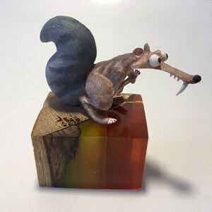 Könnte beinhalten: Eine kleine, graue und braune Eichhörnchenfigur mit großen Augen und einem zahnigen Grinsen, auf einem bunten Holzsockel mit rotem, gelbem und grünem Harz.