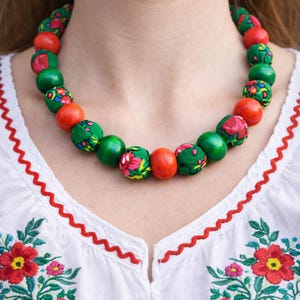 May include: A beaded necklace with alternating red and green beads. Some green beads are covered in a floral fabric. The necklace is worn over a white blouse with red embroidered floral details.
