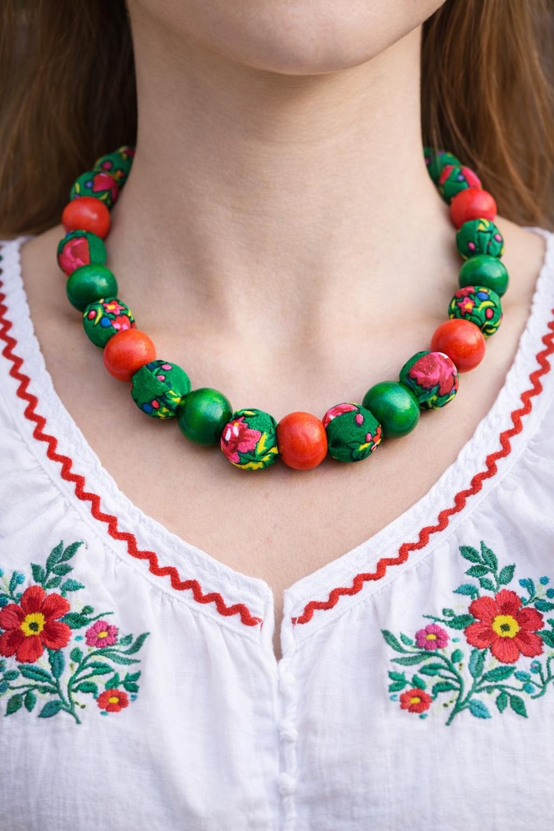 May include: A beaded necklace with alternating red and green beads. Some green beads are covered in a floral fabric. The necklace is worn over a white blouse with red embroidered floral details.