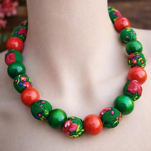 May include: A beaded necklace featuring alternating red and green spheres. Some green beads are covered in a floral fabric. The necklace is displayed on a mannequin.