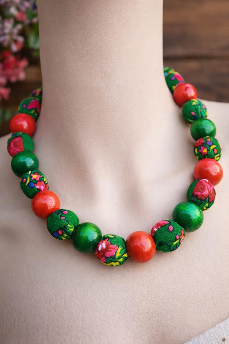 May include: A beaded necklace featuring alternating red and green spheres. Some green beads are covered in a floral fabric. The necklace is displayed on a mannequin.