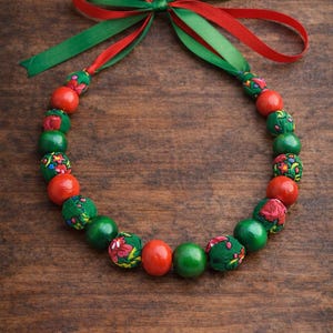 May include: A festive necklace featuring alternating red, green, and patterned fabric-covered beads. The necklace is tied with a red and green ribbon bow. The beads are approximately 1 inch in diameter.