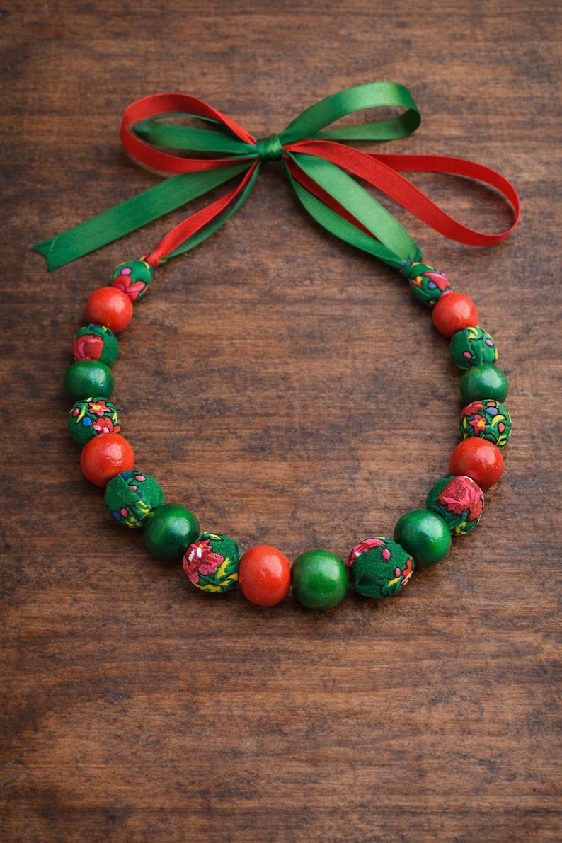 May include: A festive necklace featuring alternating red, green, and patterned fabric-covered beads. The necklace is tied with a red and green ribbon bow. The beads are approximately 1 inch in diameter.