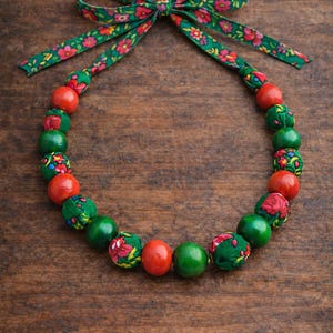 May include: A necklace with a green floral ribbon bow and a strand of alternating red, green, and fabric-covered beads. The fabric beads have a red floral pattern. The necklace is displayed on a wooden surface.