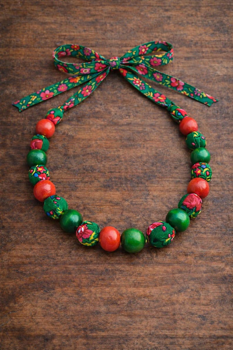 May include: A necklace with a green floral ribbon bow and a strand of alternating red, green, and fabric-covered beads. The fabric beads have a red floral pattern. The necklace is displayed on a wooden surface.