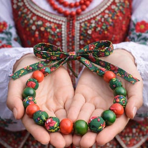 May include: A colorful beaded necklace with a green floral bow. The necklace features alternating red and green beads, some with floral patterns. The background shows traditional embroidered clothing, suggesting a cultural or folk art theme.
