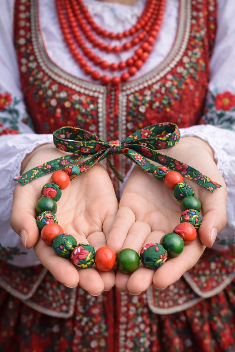 May include: A colorful beaded necklace with a green floral bow. The necklace features alternating red and green beads, some with floral patterns. The background shows traditional embroidered clothing, suggesting a cultural or folk art theme.