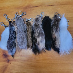 May include: A collection of keychains featuring various colors of animal fur tails. The tails are attached to silver-colored metal clasps. The fur colors range from white to black, with shades of brown and gray.