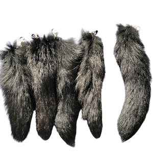 Puede incluir: Varias colas de piel sintética gris oscuro con cierres metálicos plateados. Las colas son esponjosas y varían ligeramente en forma y tamaño. Las colas están dispuestas sobre un fondo blanco.