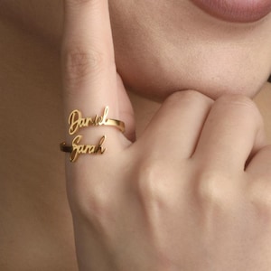 May include: A gold-coloured ring featuring the names "Daniel" and "Sarah" in a cursive font. The ring is worn on a finger, highlighting the bespoke design. The Vida Art logo is visible in the bottom right corner.