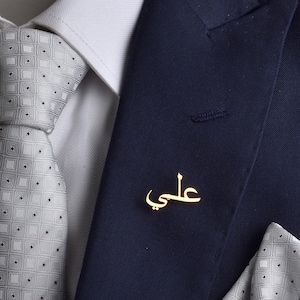May include: A navy blue suit with a white shirt, a gray patterned tie, and a matching pocket square. A gold-coloured pin with Arabic script is on the lapel. The text "THE VIDA ART" is visible.