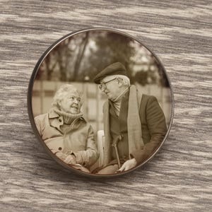 Op de afbeelding: Een ronde, sepia-kleurige pin met een ouder echtpaar dat op een bankje zit en in gesprek is. De pin heeft een zilverkleurige rand en is geplaatst tegen een achtergrond met houtnerf. Het paar draagt jassen en sjaals.