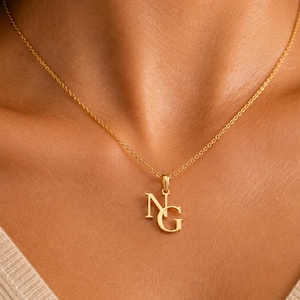 Op de afbeelding: Een goudkleurige ketting met een hanger bestaande uit de in elkaar verstrengelde hoofdletters N en G. De hanger hangt aan een fijne, dunne goudkleurige ketting. De letters zijn uitgevoerd in een klassiek schreeflettertype met een glanzende afwerking.