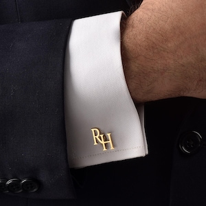 Op de afbeelding: Een close-up van een persoon in een marineblauw pak met een wit overhemd en gouden manchetknopen. De manchetknopen tonen de initialen "RH". Een gele stropdas met een stippenpatroon is zichtbaar.