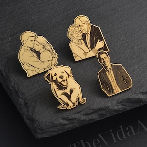 May include: A collection of gold-colored enamel pins featuring various designs. The pins depict a couple embracing, an elderly couple, a man, and a dog. The pins are arranged on a dark, textured surface.