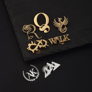 Könnte beinhalten: Eine Sammlung von Metall-Anstecknadeln, darunter goldfarbene Designs des Buchstabens Q, einer Rose, eines Phönix, eines Unendlichkeitssymbols mit Stadtsilhouetten und des Wortes WALK. Zwei silberfarbene Anstecker zeigen die Buchstaben AK im Kranz.