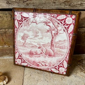 May include: Square framed artwork with a red and white pastoral scene. The image shows two sheep under a tree, with a fence and hills in the background. The frame is made of dark brown wood. The artwork is a decorative piece.