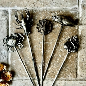 May include: Set of six silver-toned metal cocktail picks with different sea creature designs. The picks feature a crab, a lobster, a fish, a swan, and two different leaf designs.