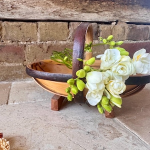 Könnte beinhalten: Ein Holzkorb mit Griff, gefüllt mit weißen Blumen und grünen Knospen. Der Korb hat einen dunkelbraunen Rand und Griff und steht auf einem kleinen Holzständer. Der Hintergrund ist eine Backsteinmauer.