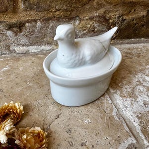 以下が含まれることがあります： 白いセラミック製のアヒルの置物。楕円形の皿の上に置かれ、アヒルは質感のある表面を持ち、皿の中に収まっています。石の表面に置かれ、手前にドライフラワーが飾られています。