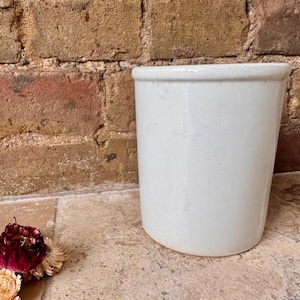 May include: A cylindrical, off-white ceramic container with a slightly flared rim. The container has a speckled texture and sits on a stone surface. The background features a weathered brick wall, and dried flowers are in the foreground.