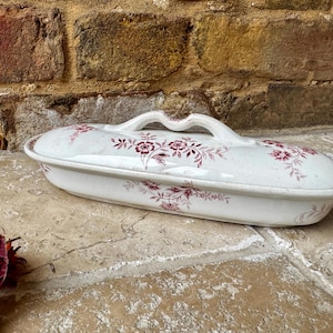 Peut inclure: Plat de service ovale en céramique blanche avec couvercle, orné de motifs floraux bordeaux et d'une poignée. Le plat repose sur une surface en pierre, avec un mur de briques en arrière-plan. Environ 30 cm de long.