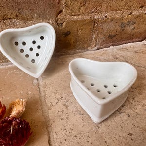 May include: Two white heart-shaped ceramic soap dishes with perforated bases. Each dish features a smooth, glossy finish and a series of circular drainage holes arranged in a pattern across the bottom, designed to hold a bar of soap.