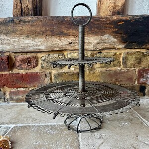 Könnte beinhalten: Zweistöckiger Servierständer im antiken Stil aus Draht. Der Ständer hat einen kreisförmigen Sockel und zwei kreisförmige Etagen, die durch eine zentrale Stange mit Griff verbunden sind. Der Draht hat eine dunkle, gealterte Oberfläche.