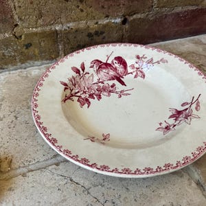 May include: Cream-coloured ceramic plate with a maroon floral and bird design. The plate has a scalloped edge with a repeating maroon floral pattern. The centre of the plate features two birds perched on a branch.