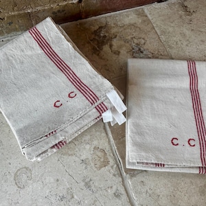 Può includere: Due asciugamani di lino piegati. Gli asciugamani sono bianco sporco con tre strisce rosse e ricamati con le lettere "C.C.". Gli asciugamani hanno etichette bianche attaccate. Gli asciugamani sono su una superficie di pietra.