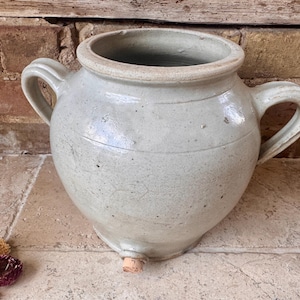 May include: A light grey glazed ceramic vessel with a wide circular opening, two side handles, and a small cork stopper at the base. The surface features a subtle crackled texture and minor dark speckling throughout the rounded body of the pot.