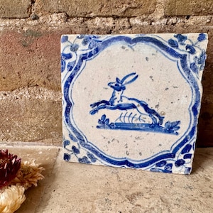 Può includere: Piastrella quadrata in ceramica con un design blu e bianco. La piastrella presenta una lepre che salta al centro, circondata da un bordo floreale blu. La piastrella è probabilmente vintage o antica.