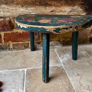 May include: A small, antique wooden stool with a teal painted finish and floral design on the seat. The stool has three legs and shows signs of wear, adding to its vintage charm. The background features a brick wall and stone floor.