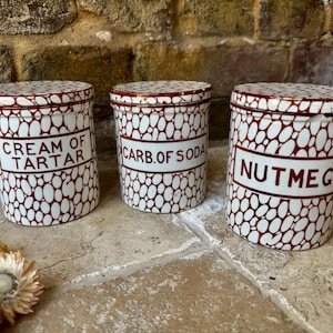 May include: Three white ceramic canisters with red and white speckled patterns. The canisters have white labels with the words "Cream of Tartar", "Carb. of Soda", and "Nutmeg" printed in black.