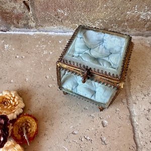 Puede incluir: Un joyero cuadrado de cristal con un marco metálico texturizado en tono latón y una tapa con bisagras. El interior está forrado con un cojín de tela azul claro acolchado. La caja cuenta con un pequeño cierre decorativo de metal.